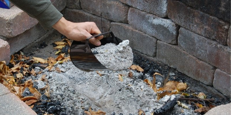 A person's hand using a metal scoop to clean out ash and debris from a stone fire pit, with remnants of burnt wood and fallen leaves around